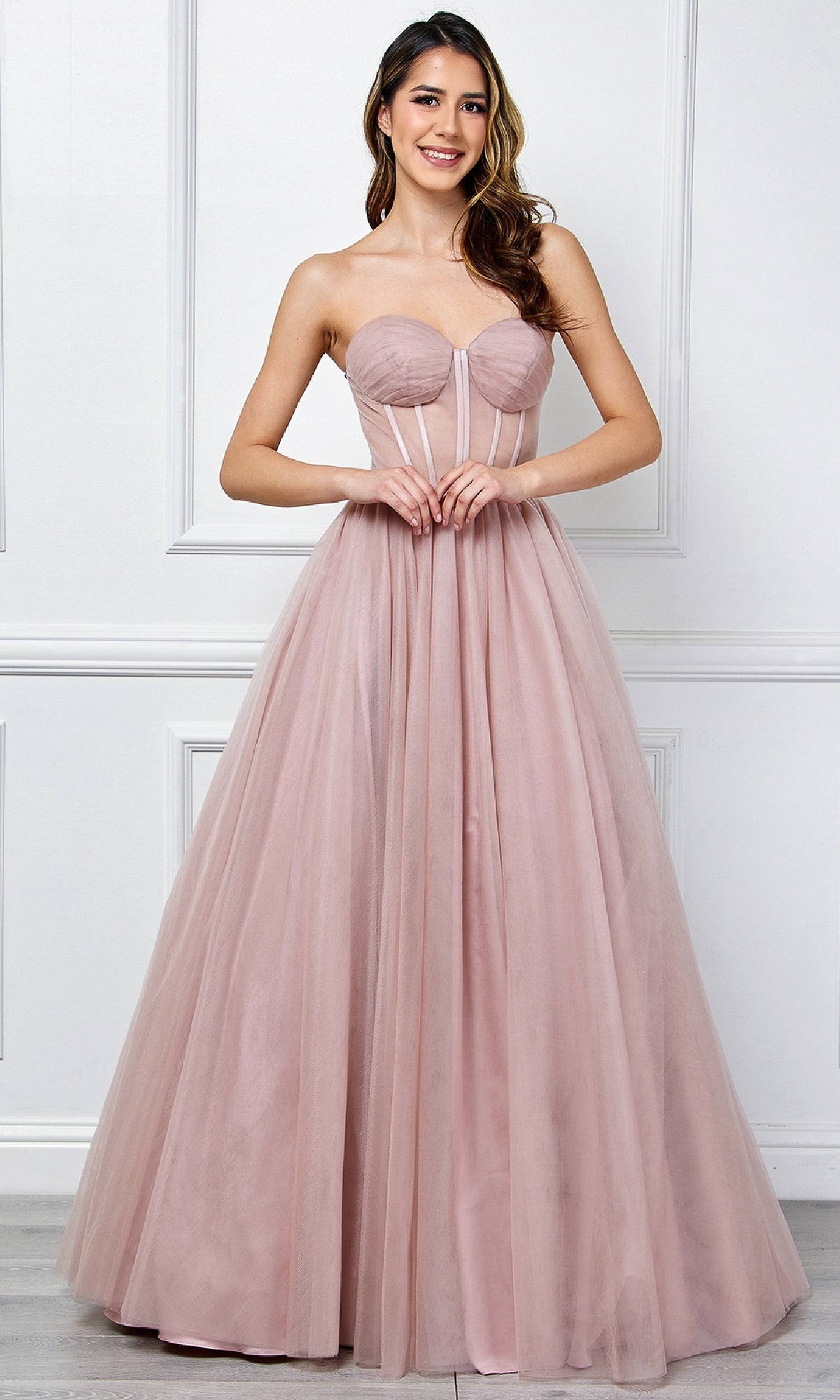 A woman poses elegantly against a white paneled wall in the Strapless Corset Prom Ball Gown: Amelia Couture 6202โa blush pink tulle ball gown featuring a fitted bodice and full skirt.